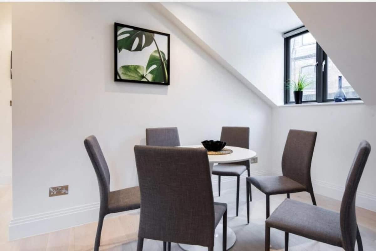 A dining area with a round table and four chairs, a black bowl on the table, and a framed plant on the wall.