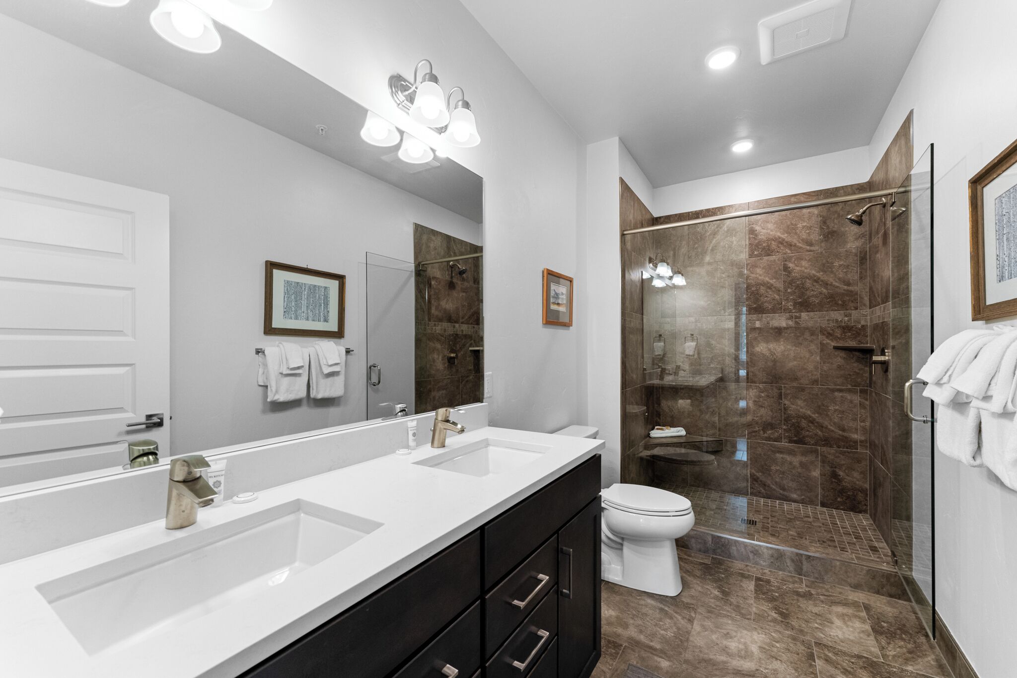 A modern bathroom with a large mirror, two sinks, a toilet, and a glass-enclosed shower.
