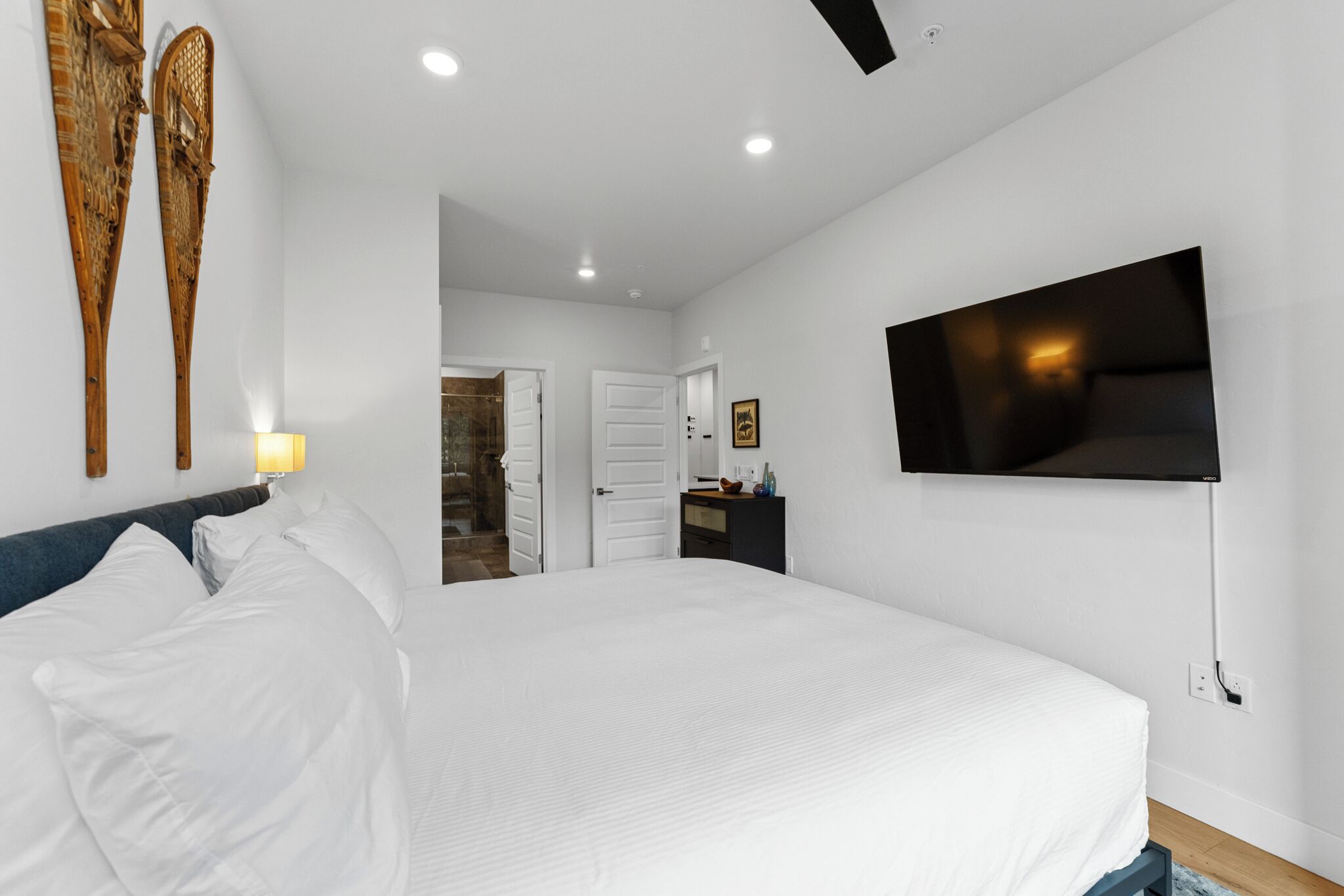 A modern hotel room with a large bed, a flat-screen TV mounted on the wall, and a small desk with a lamp.