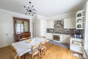 A kitchen with white cabinets, a dining table with chairs, a chandelier, and a wooden cabinet with glass doors.