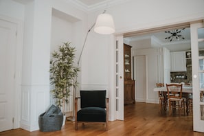 A room with a wooden floor, a dark chair, a lamp, a plant, and a dining area with a table and chairs.