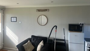 A living room with a black sofa, a vacuum cleaner, a microwave, and a refrigerator. - Chalet for Hire in Florida Hemsby (Great Yarmouth)