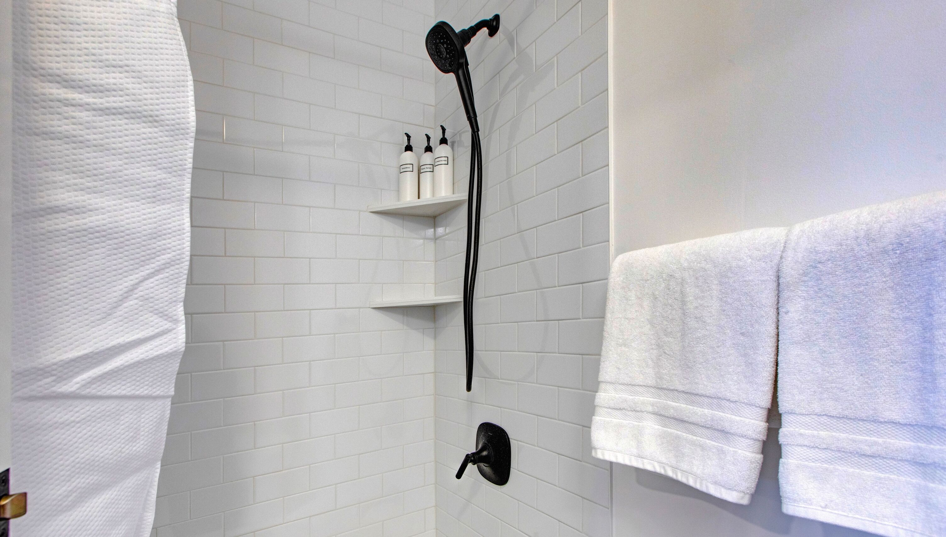 A bathroom with white tiled walls, a black showerhead, and two shelves holding toiletries.