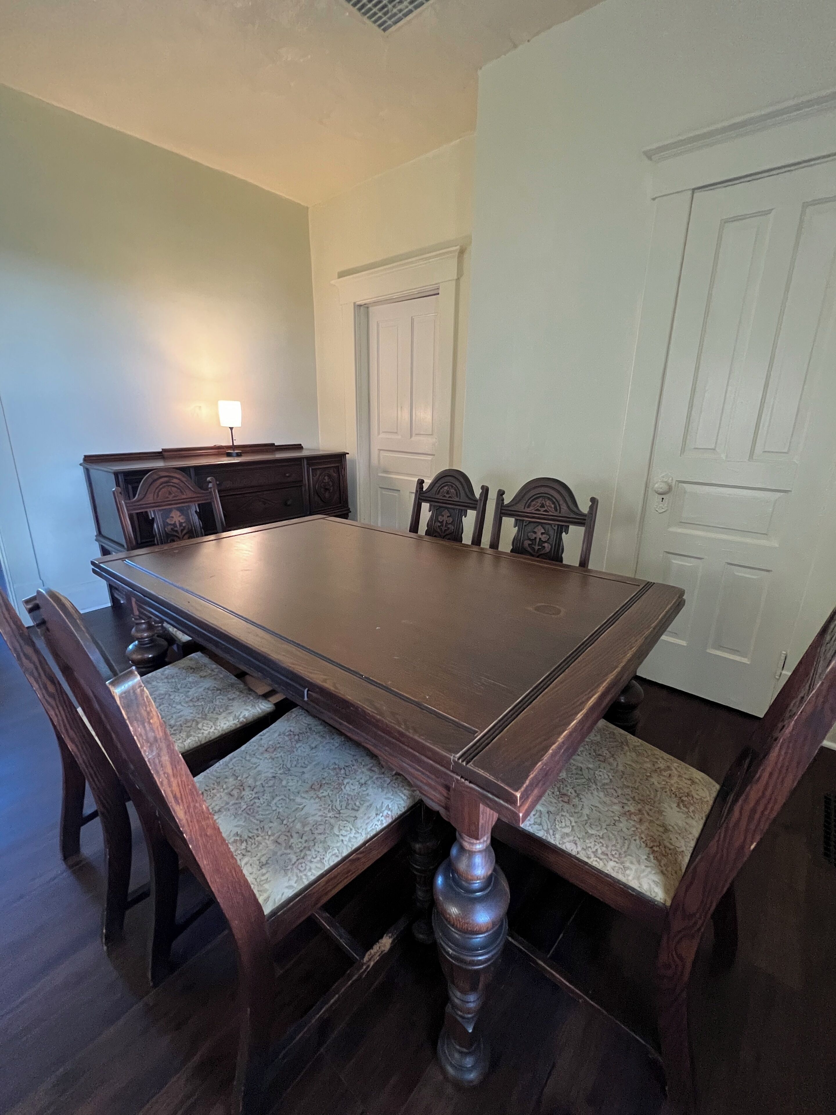 Uma sala de jantar com uma grande mesa de madeira, seis cadeiras, um aparador e uma porta.