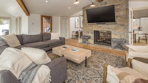 A living room with a stone fireplace, a large flat-screen TV mounted on the wall, and a cozy seating area with a sofa and armchairs. - Luxury 7 Bedroom Home + Hot Tub + Views of Lone Peak (Big Sky)