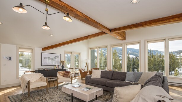 A spacious living room with a large gray sofa, a white coffee table, and a patterned rug. There are multiple windows offering a view of a snowy landscape. - Luxury 7 Bedroom Home + Hot Tub + Views of Lone Peak (Big Sky)