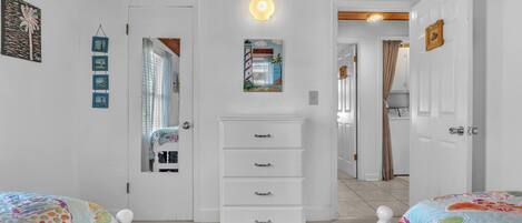 Un dormitorio con un tocador blanco, una cama con colcha de colores, una lámpara de techo y una puerta que da a otra habitación.