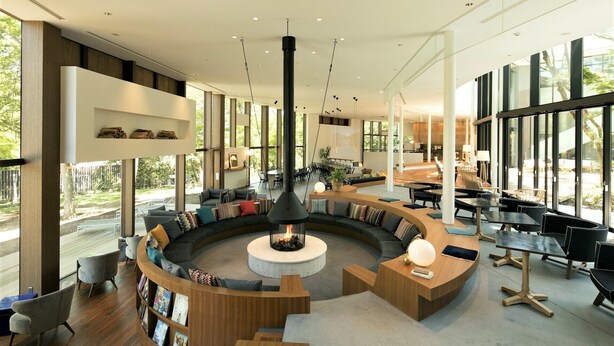 Un espace de détente moderne avec un aménagement circulaire central, une cheminée et de grandes fenêtres offrant une vue sur l’extérieur.