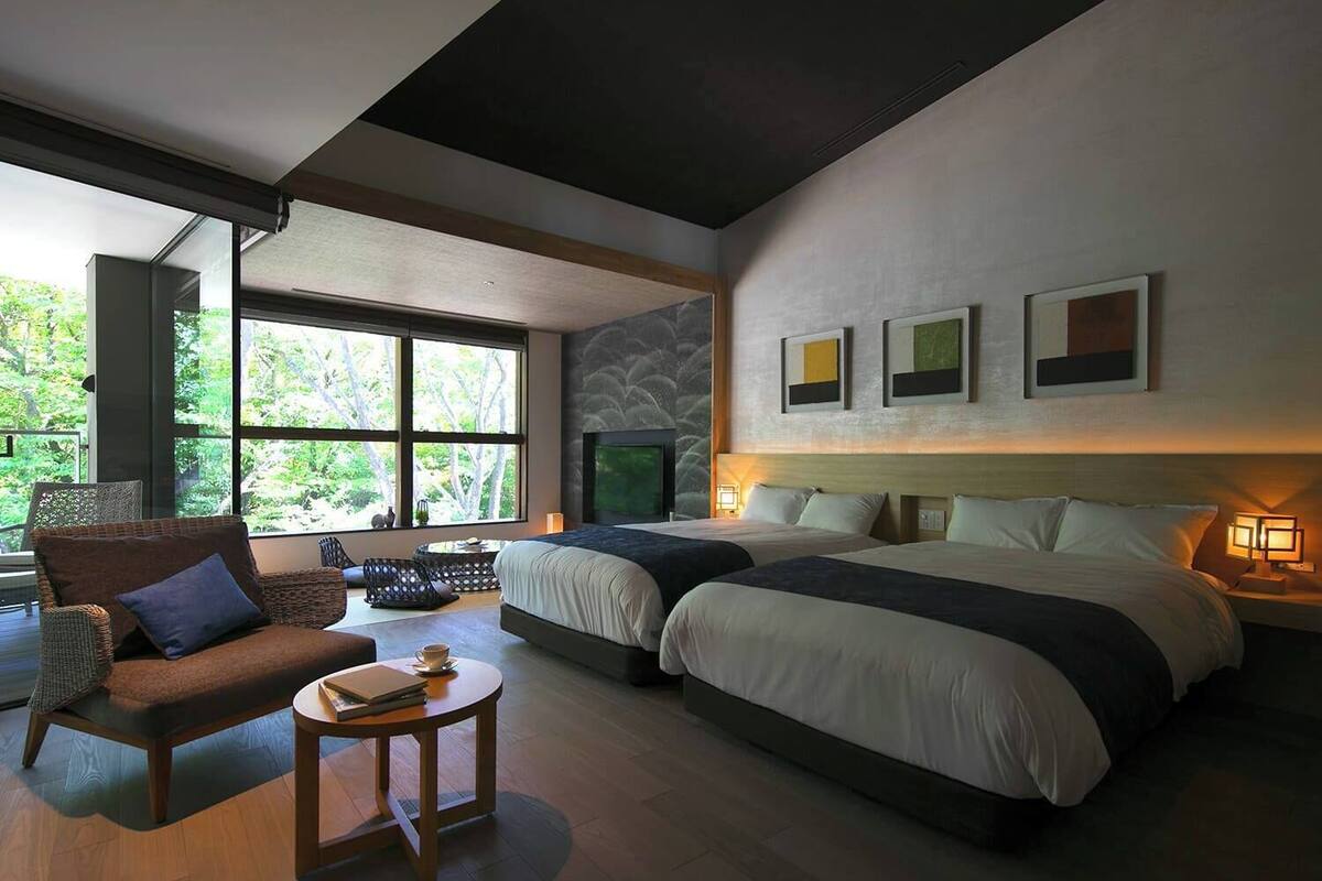 Habitación de hotel moderna con dos camas, un sofá, una mesita y un ventanal con vistas a árboles.