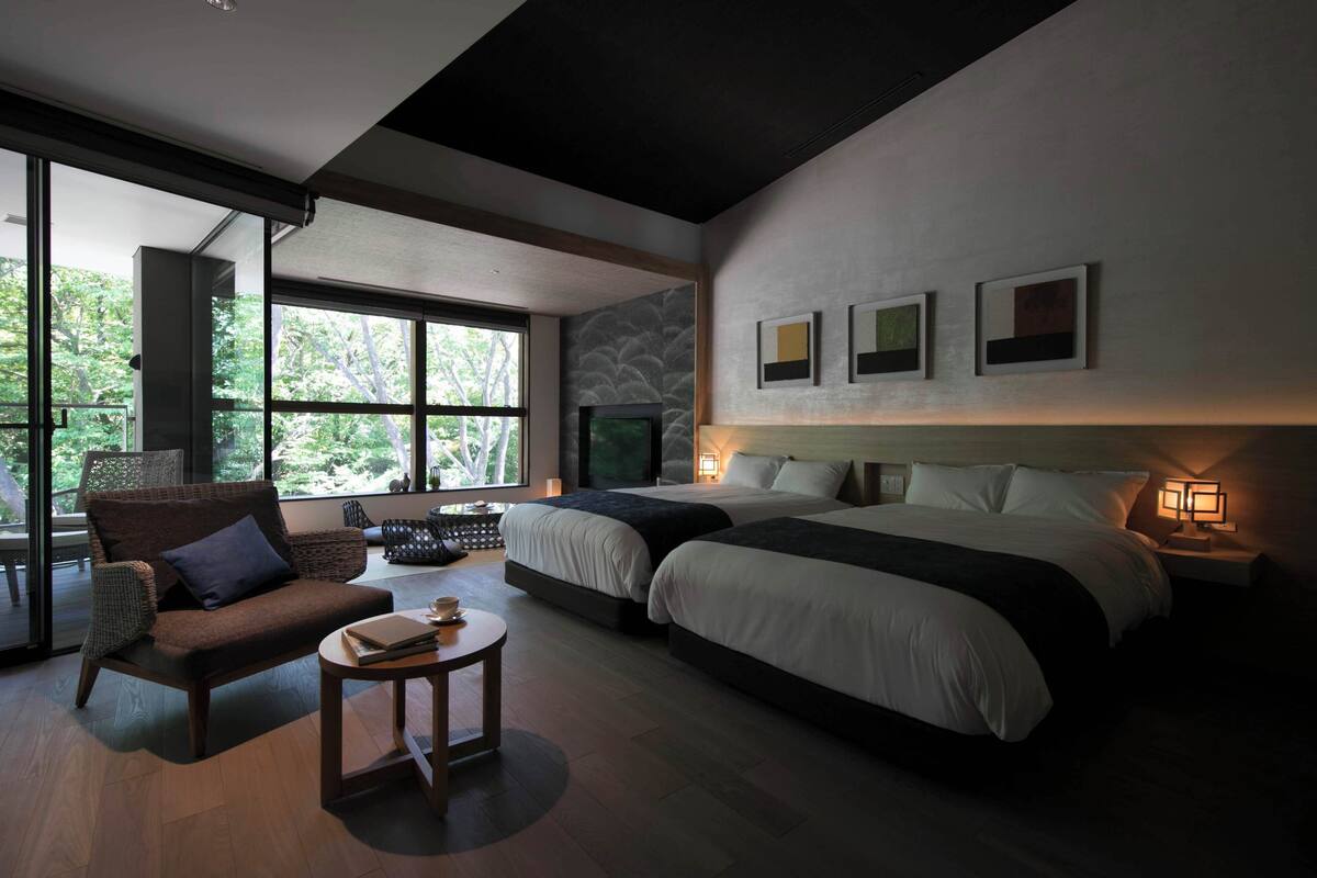 Una habitación de hotel moderna con dos camas, un sofá, una mesita y ventanas amplias.