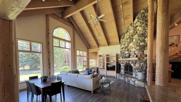 A spacious living area with a stone fireplace, wooden ceiling beams, and large windows offering views of the outdoors. - Luxurious Log Home Retreat (Hailey)