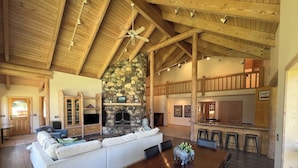 A spacious living area with a stone fireplace, wooden ceiling beams, and a large dining table with bar stools. - Luxurious Log Home Retreat (Hailey)