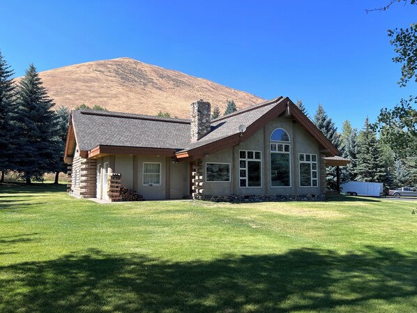 A log cabin with a stone chimney, large windows, and a mountain backdrop.