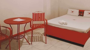 Ein kleines Bett mit rotem Rahmen, weiße Bettwäsche und Kissen, ein kleiner roter Tisch und ein roter Sessel in einem Raum mit weißen Wänden und rotem Fußleistenverlauf.