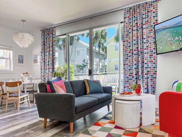 A modern living room with a gray sofa, wooden chairs, a white coffee table, and colorful curtains.