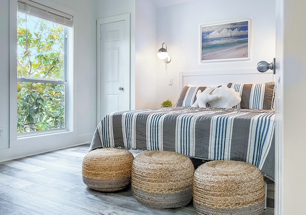 A bedroom with a large window, a bed with striped bedding, and three woven ottomans.