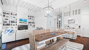 Amplia sala de estar con sofá blanco, mesa de comedor de madera, sillas de mimbre y televisor montado en la pared.