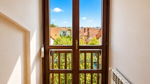 A wooden door with a glass panel, a balcony with a metal railing, and a view of buildings and greenery.