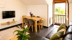 A modern living room with a flat-screen TV, a wooden dining table with chairs, a sofa, and a large window with a view of greenery.