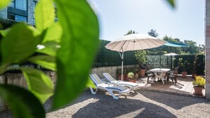A patio area with two sun loungers and a table under a large umbrella, surrounded by potted plants and a hedge.