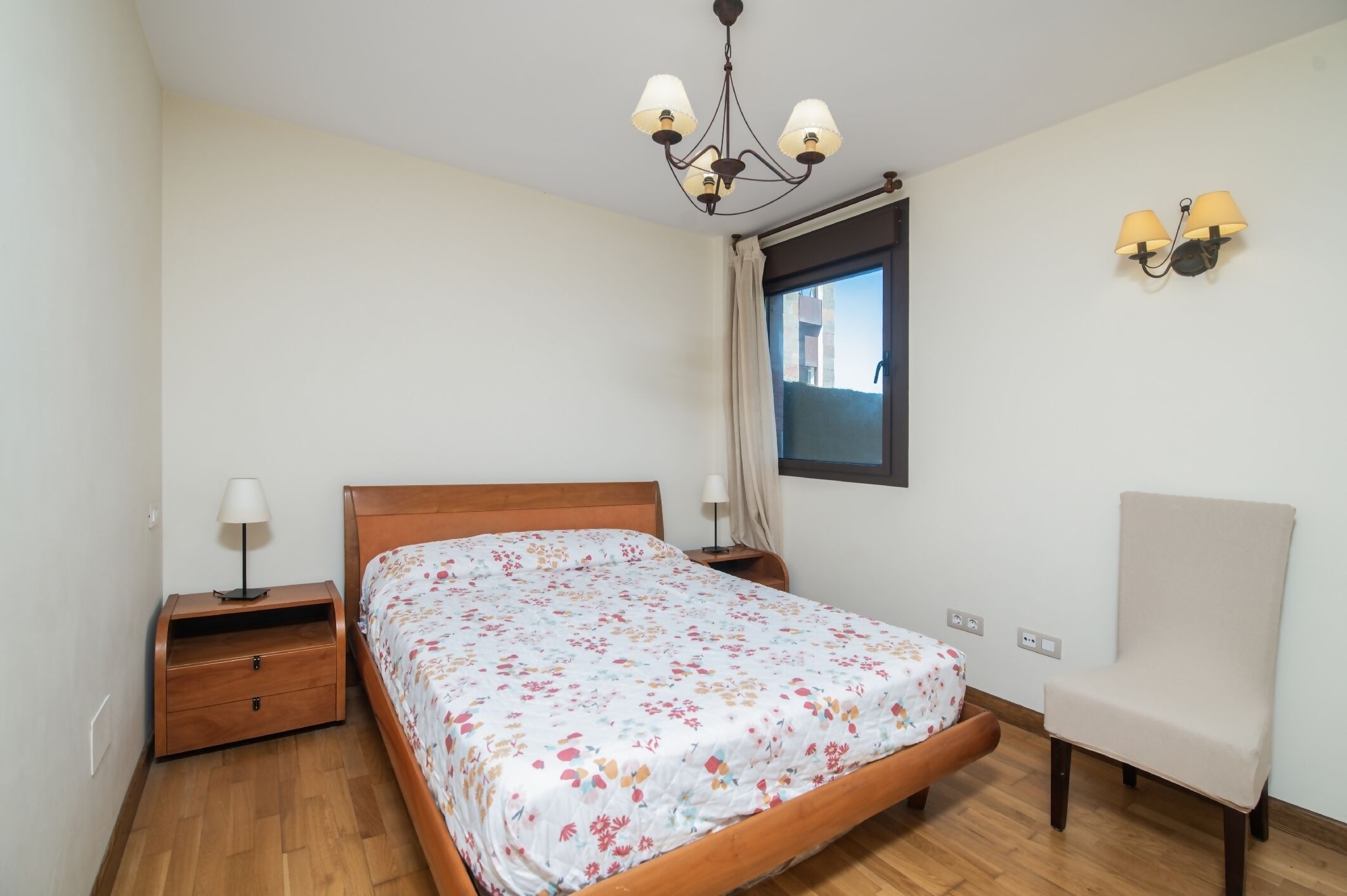 A bedroom with a wooden bed, a nightstand, a chair, a window with curtains, and a chandelier.