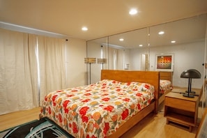 A bedroom with a wooden bed, a nightstand, a lamp, a mirror, and a window with curtains. - Stunning Oceanfront Venice Beach 2BR/2BA with Beach Views + Free Parking (Los Angeles)
