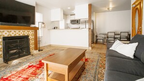 A living room with a flat-screen TV, a fireplace, a wooden coffee table, a grey sofa, and a kitchen in the background. - Stunning Oceanfront Venice Beach 2BR/2BA with Beach Views + Free Parking (Los Angeles)