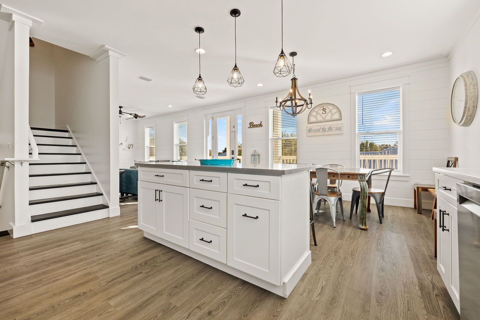 A modern kitchen with white cabinetry, a central island, pendant lighting, a dining area with a table and chairs, and a staircase leading to another level.