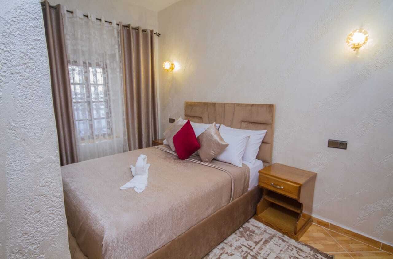 Una cama bien hecha con una colcha color beige y almohadas, una mesita de noche de madera con un cajón, y una ventana con cortinas.