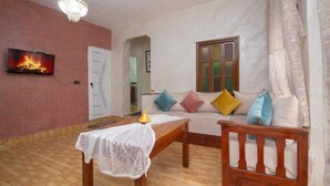 Una sala de estar con pared de ladrillo, un televisor montado sobre una chimenea, un sofá con cojines coloridos, una mesa de centro de madera y una araña.