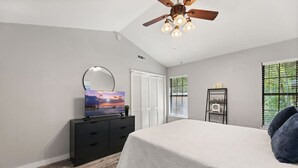 A bedroom with a bed, a dresser, a TV, a ceiling fan, and a window with blinds. - The Pine Grove Retreat (Ladson)