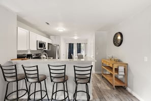 Una cocina moderna con armarios blancos, una isla central con taburetes de bar, un microondas y una mesita auxiliar de madera.
