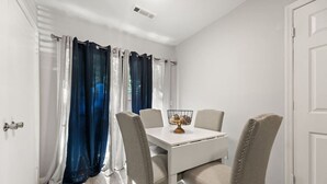 A dining area with a white table, four upholstered chairs, a metal bowl on the table, and a window with blue and white curtains. - The Pine Grove Retreat (Ladson)