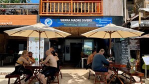 Een terras met houten tafels en parasols, een bord voor Sierra Madre Divers, en uitzicht op de oceaan.