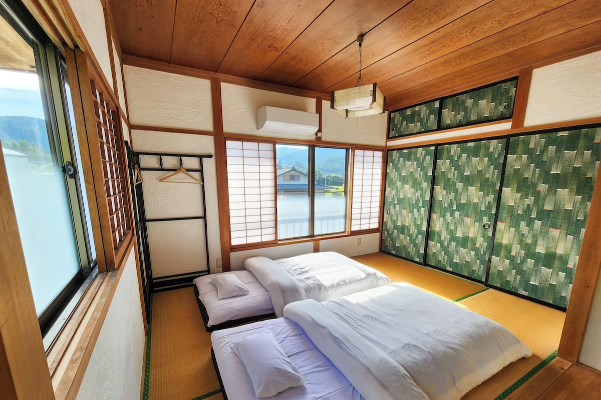 Una habitación tradicional japonesa con suelo de tatami, dos camas con ropa blanca, un celosía japonés y vista a la montaña.