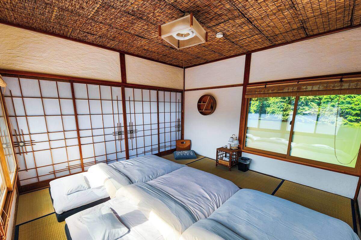Una habitación japonesa tradicional con suelo de tatami, un futón, una pequeña mesa de madera con una lámpara y una ventana con vistas a la vegetación.