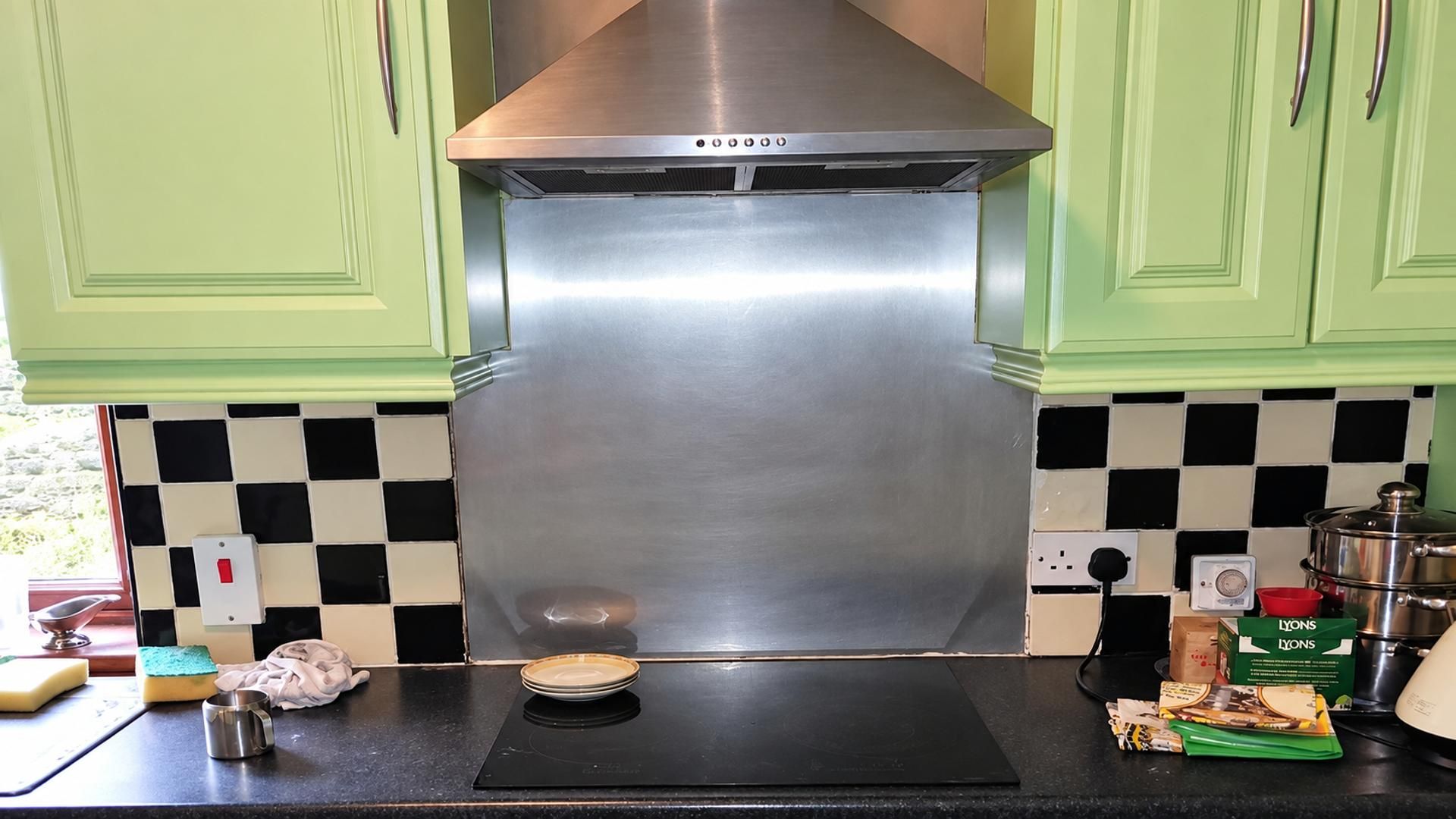 A kitchen with green cabinets, a stainless steel range hood, a black countertop, and a checkered backsplash.