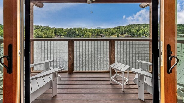 A wooden deck with two white tables and benches, a ceiling fan, and a view of a lake with houses and trees in the background. - Sleep Lakeside in Our Cozy Boathouse with Mtn Views (Landrum)
