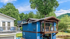 A blue houseboat with a wooden deck and a balcony, floating on a body of water. - Sleep Lakeside in Our Cozy Boathouse with Mtn Views (Landrum)