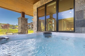 Un bain à remous installé sous une pergola en bois, avec des murs en pierre et de grandes fenêtres.