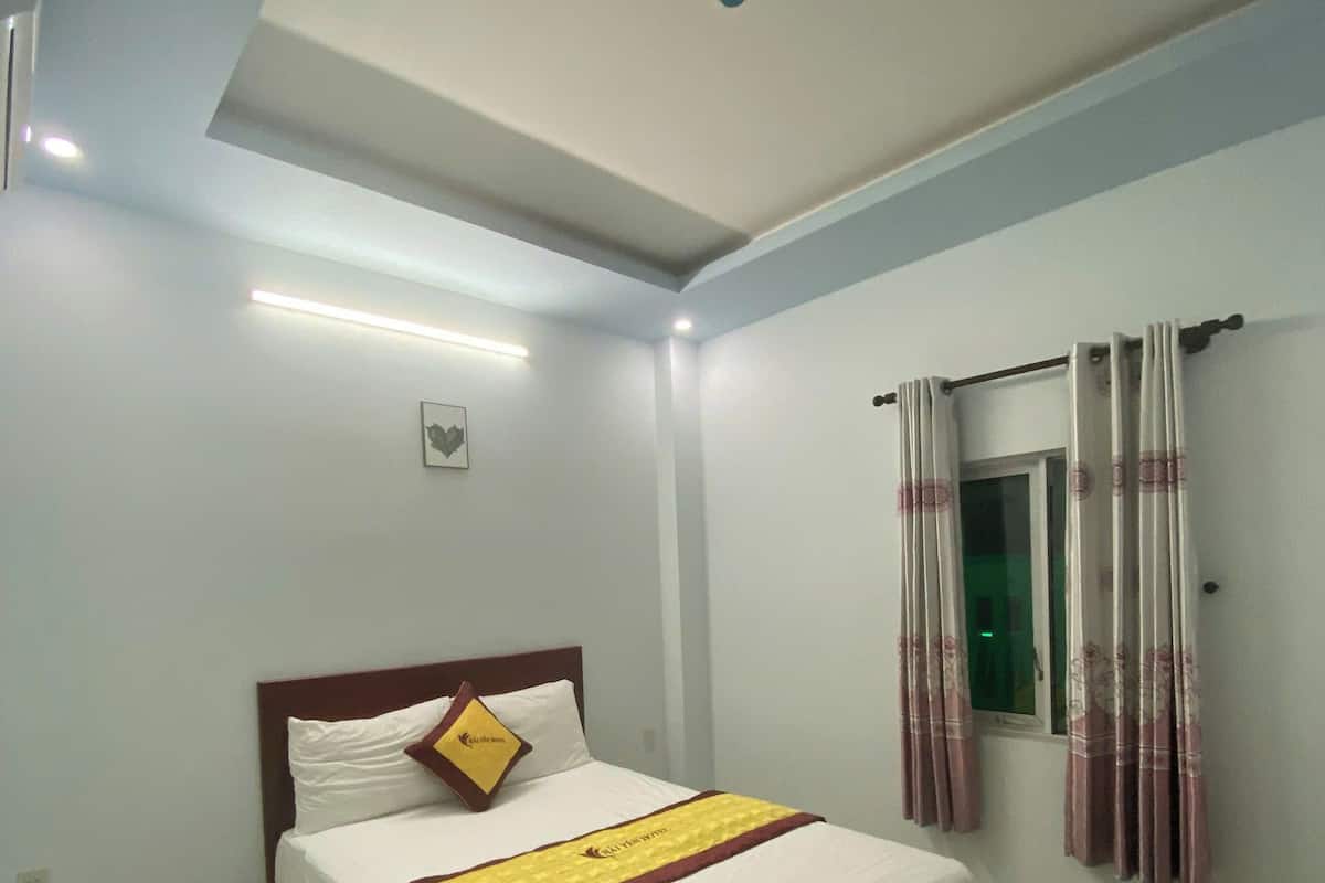 Una cama bien hecha con colcha blanca y amarilla, cabecera de madera y una sola almohada con un logotipo.