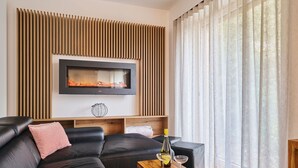 Un salon moderne avec un canapé en cuir noir, une table basse en bois sur laquelle se trouvent une bouteille et des verres, un téléviseur à écran plat fixé sur un panneau en bois, et des rideaux légers à une fenêtre.