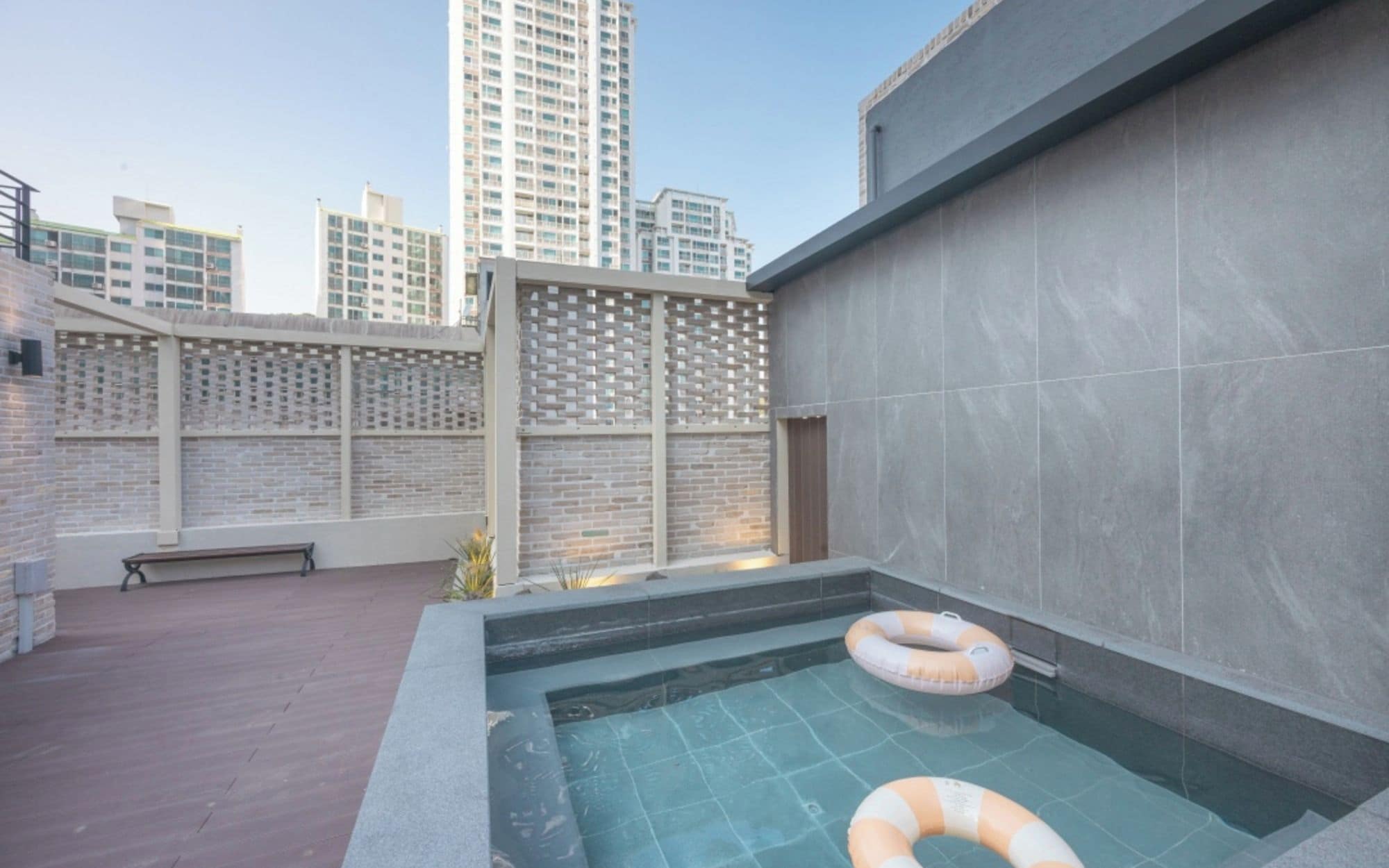 A rooftop swimming pool with a bench and a lifebuoy, surrounded by a brick wall and a high-rise building in the background.