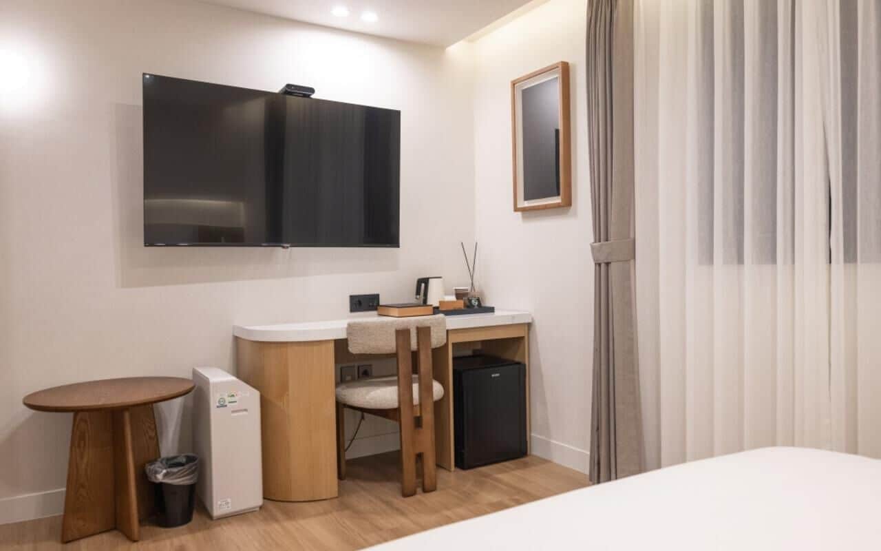 A compact hotel room with a bed, a small desk, a chair, a TV mounted on the wall, a mini-fridge, a trash bin, and a window with curtains.