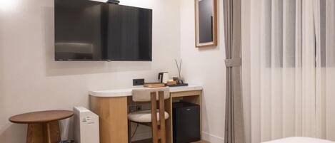 A compact hotel room with a bed, a small desk, a chair, a TV mounted on the wall, a mini-fridge, a trash bin, and a window with curtains.