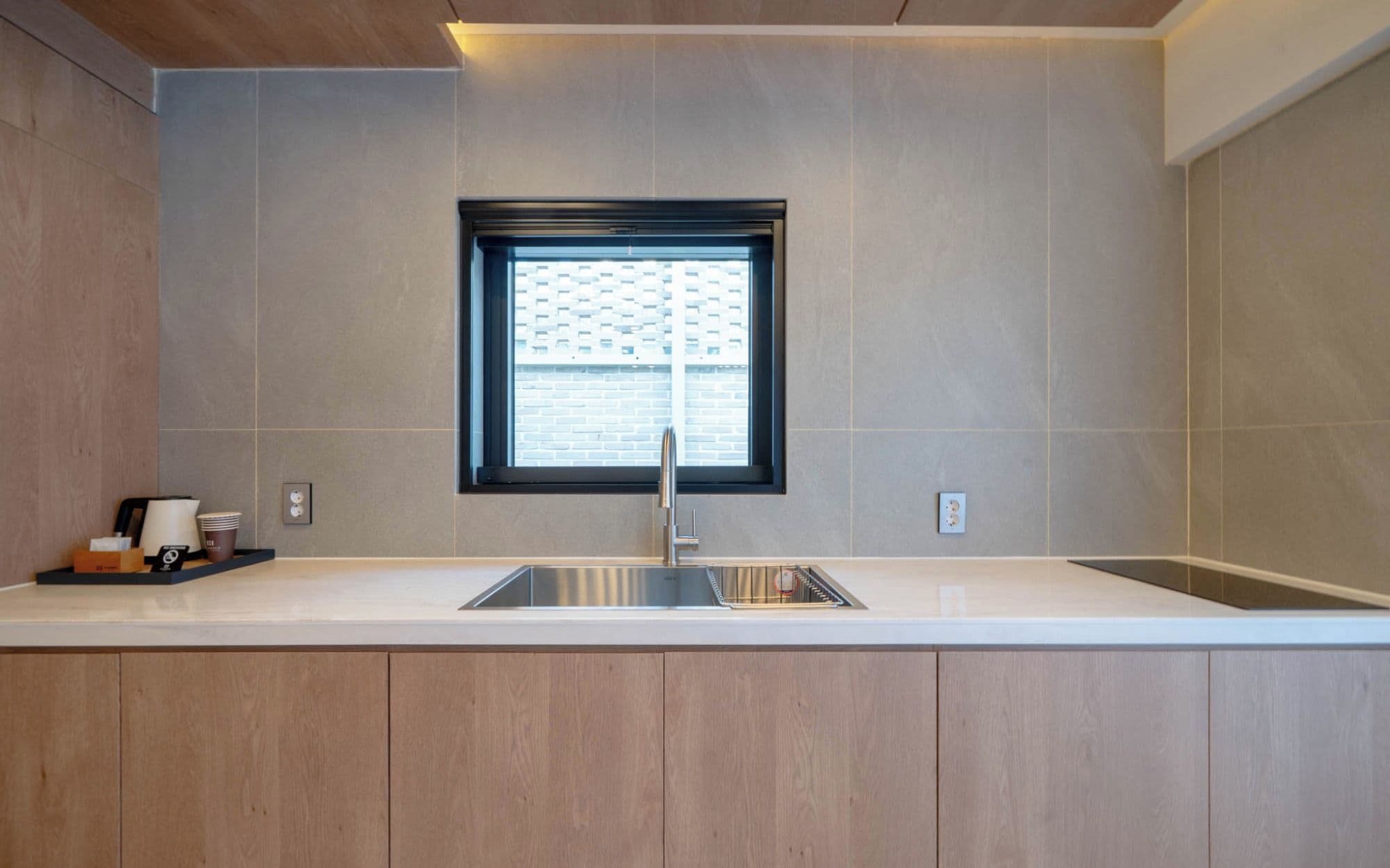 A modern kitchen with a built-in sink, a window with a view of a brick building, and a countertop with a coffee maker and cups.