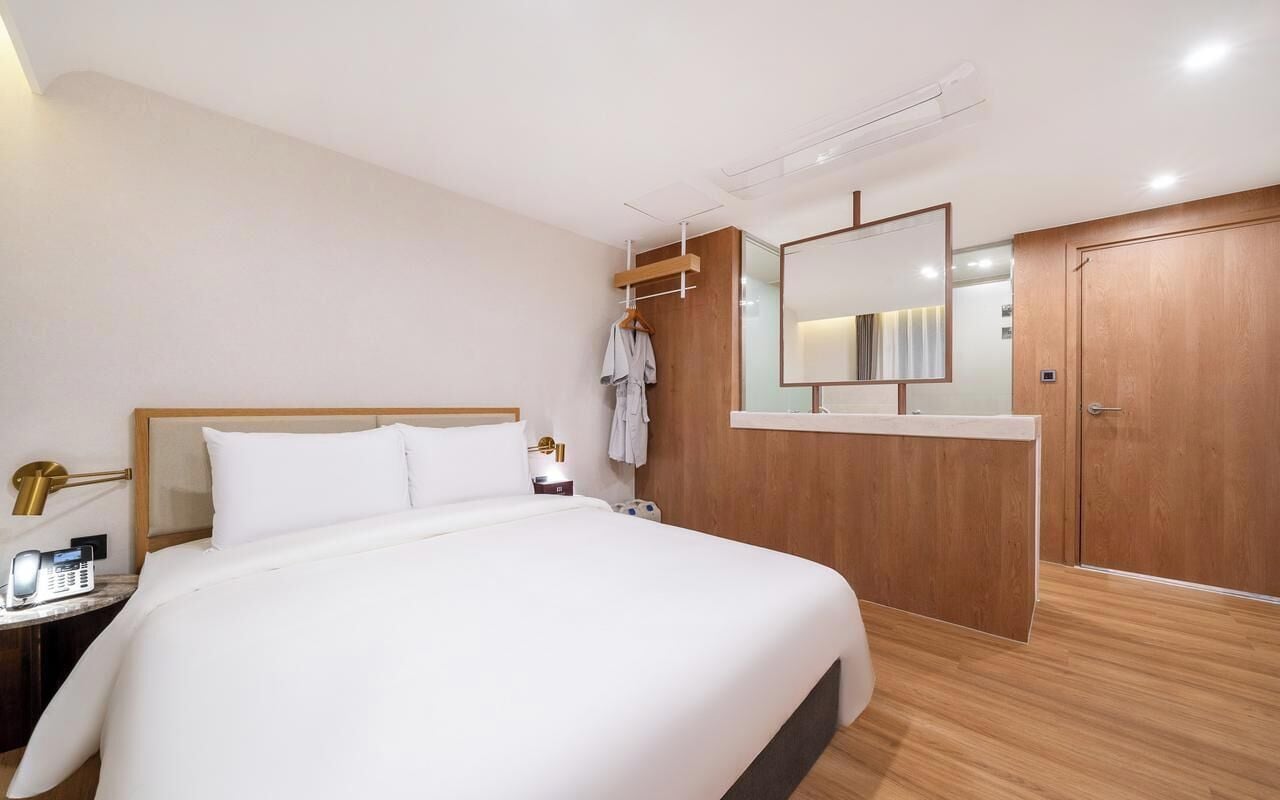 A hotel room with a large bed, a bedside table with a lamp, a mirror, a wooden wardrobe, and a robe hanging on a rack.
