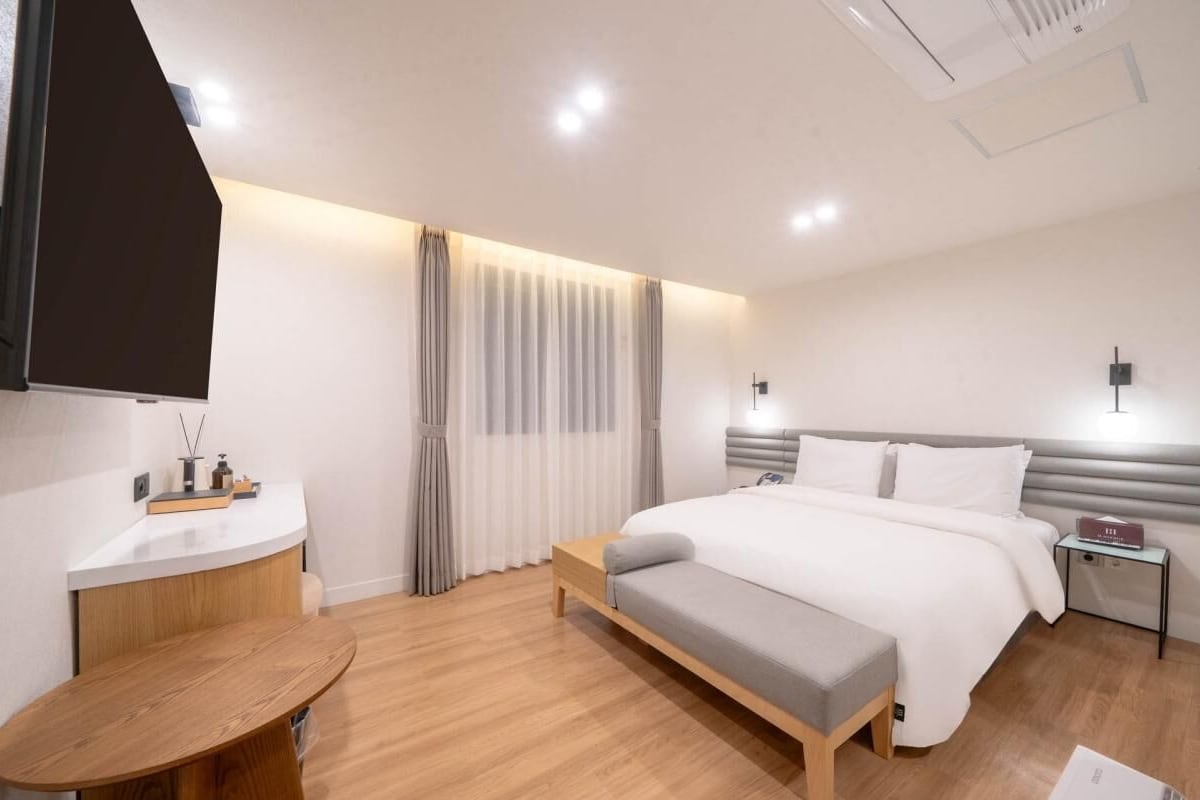 A modern hotel room with a bed, bedside table, bench, and a small table with a chair.