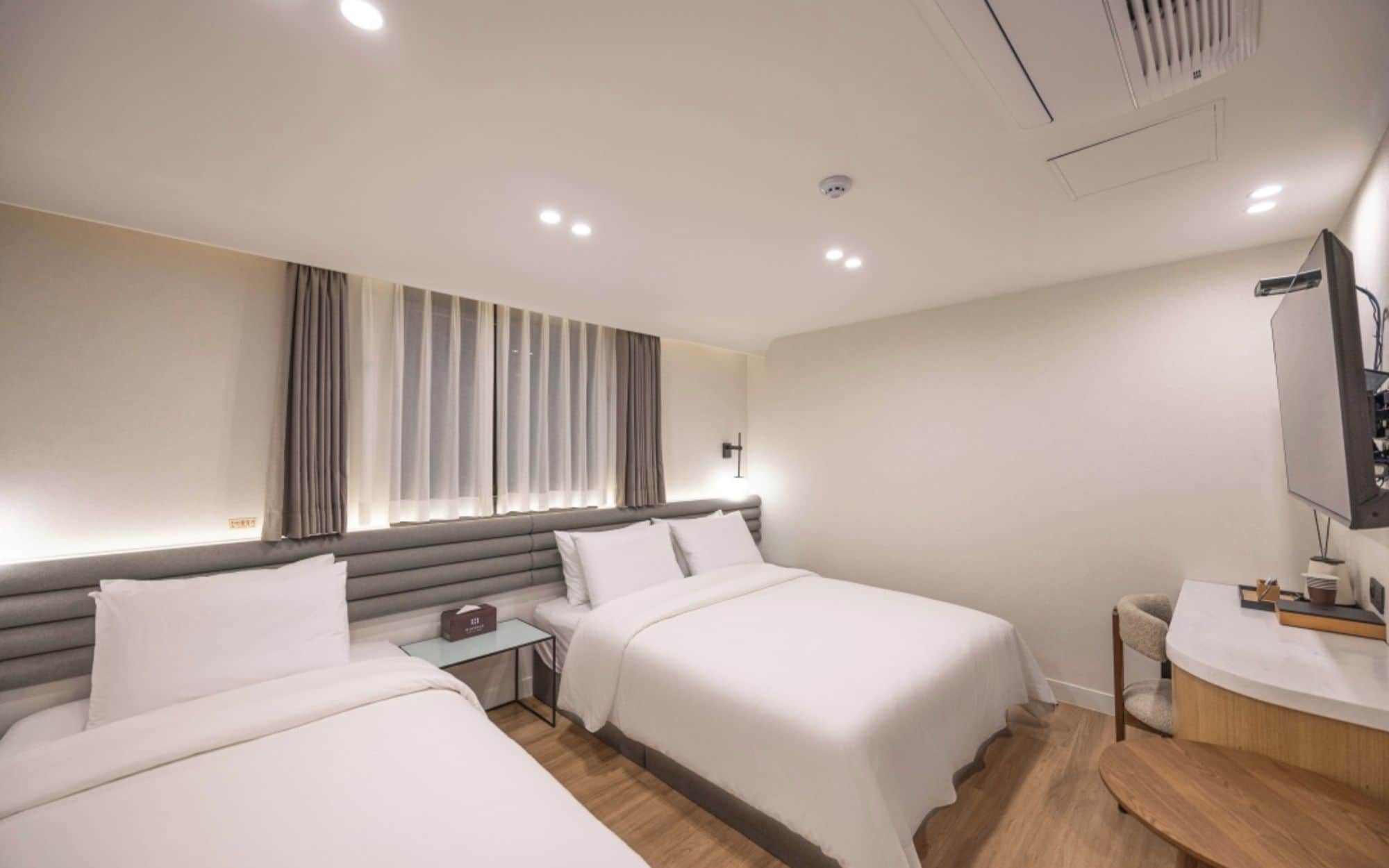 Quarto de hotel com duas camas, mesa de cabeceira, escrivaninha, cadeira, televisão e espelho.