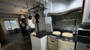 Eine Küche mit Kochinsel, Herd und hängenden Töpfen. Ein Fenster mit Jalousien, ein Kühlschrank und eine Mikrowelle sind ebenfalls vorhanden.